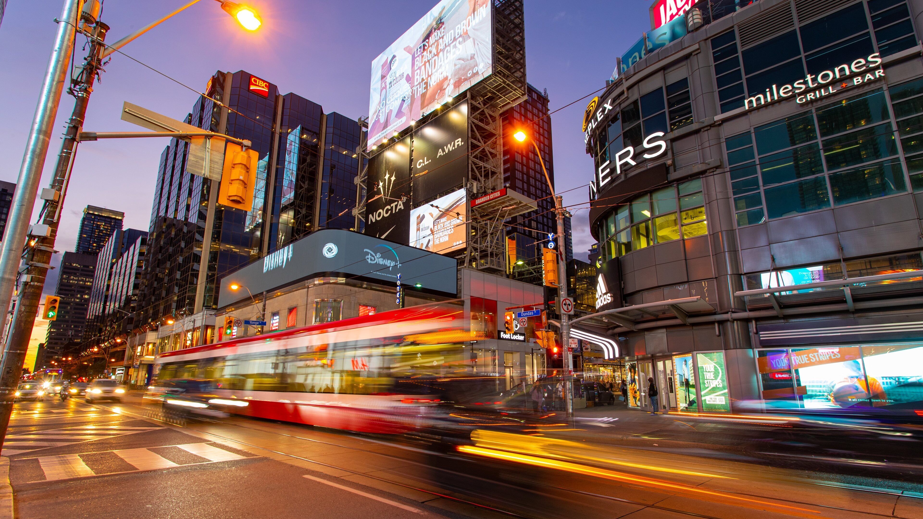 A busy urban street with a tram, illuminated billboards, and various commercial buildings.
