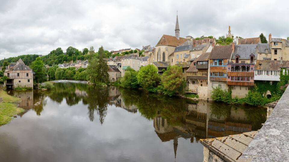 A river reflecting buildings, a church steeple, and lush greenery in a town.