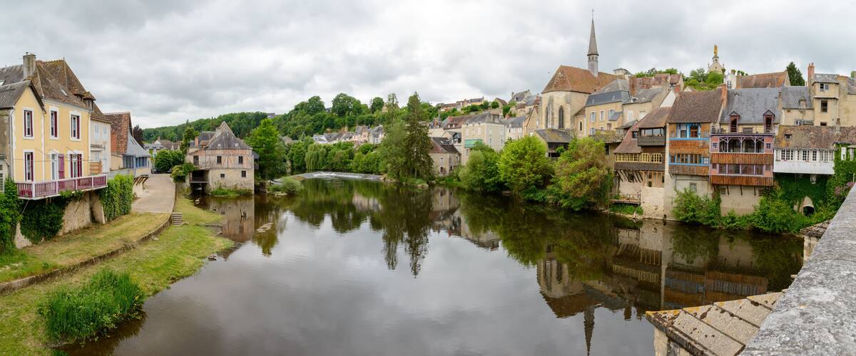 A river reflecting buildings, a church steeple, and lush greenery in a town.