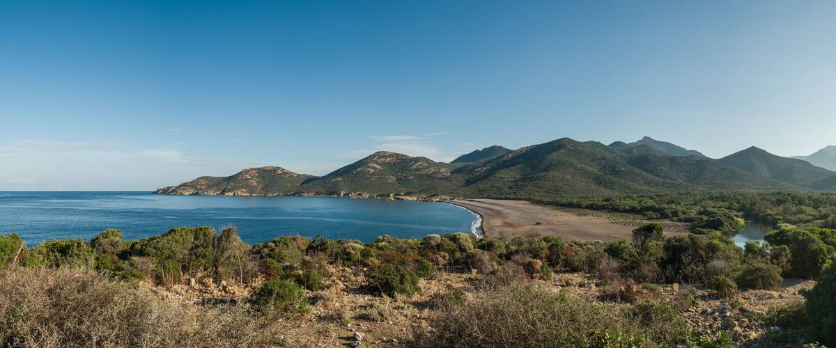 A coastal landscape with a beach, mountains, and dense vegetation.