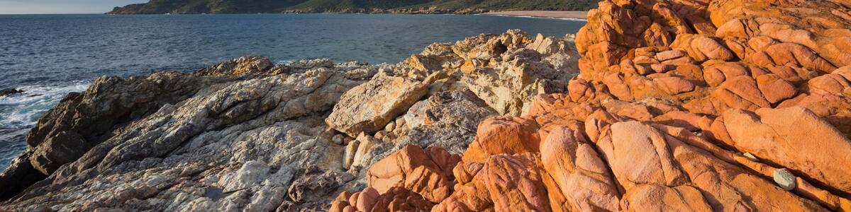 A rocky coastline with red and white formations, a calm sea, and green hills in the background.