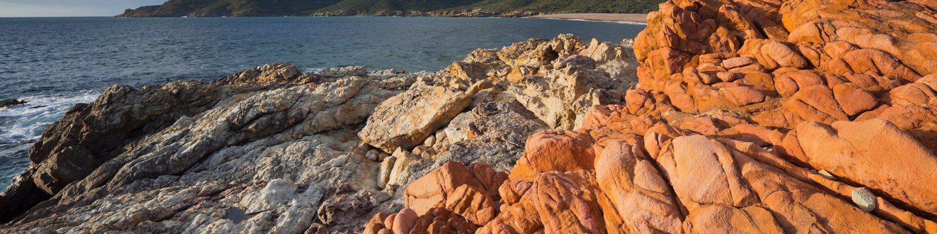 Una costa rocosa con formaciones rojas y blancas, un mar tranquilo y colinas verdes al fondo.