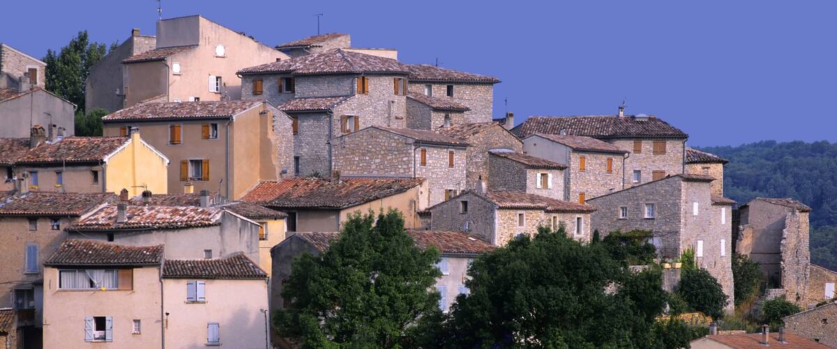 A cluster of stone buildings with terracotta roofs, surrounded by trees and set against a clear blue sky.