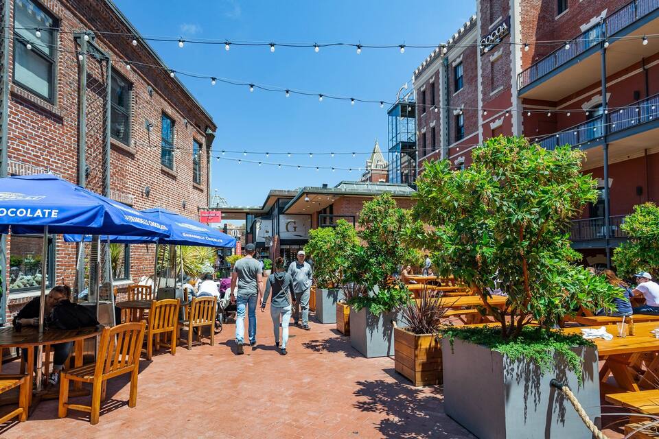 A sunny outdoor cafe with brick buildings, blue umbrellas, and wooden tables.