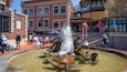 A fountain with two turtles and water jets in a public square with brick buildings and people around.