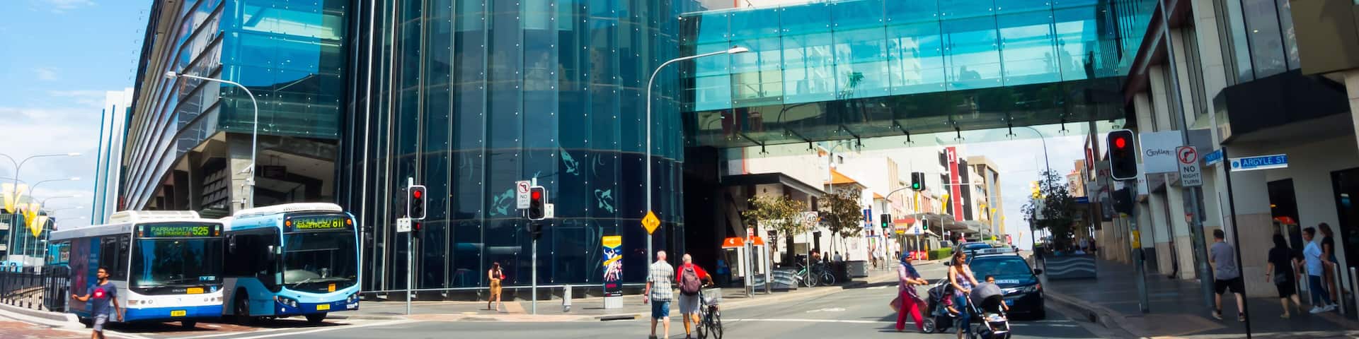 Un edificio moderno con fachada de vidrio curvada y sección superior roja, al lado de un paso peatonal y una calle concurrida con vehículos y peatones.