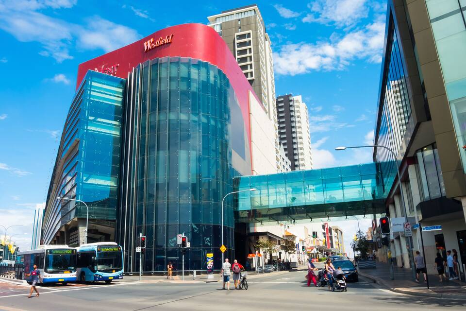 Un bâtiment moderne doté d’une façade en verre incurvée et d’une partie supérieure rouge, situé à côté d’un passage piétonnier et d’une rue animée où circulent des véhicules et des piétons.