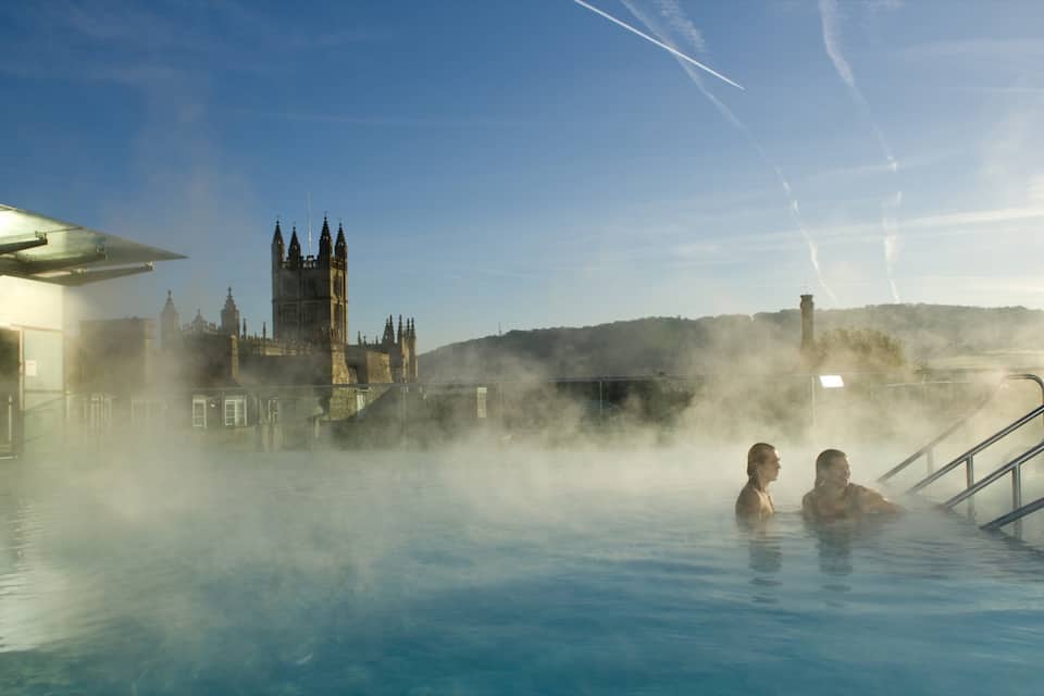 A hot spring pool with steam rising, a historic building in the background, and a clear sky.