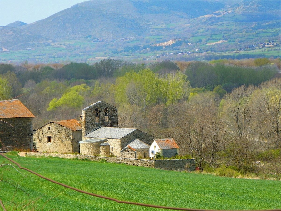 Ländliche Landschaft mit Steinhäusern, einer Kirche und sanften Hügeln im Hintergrund.