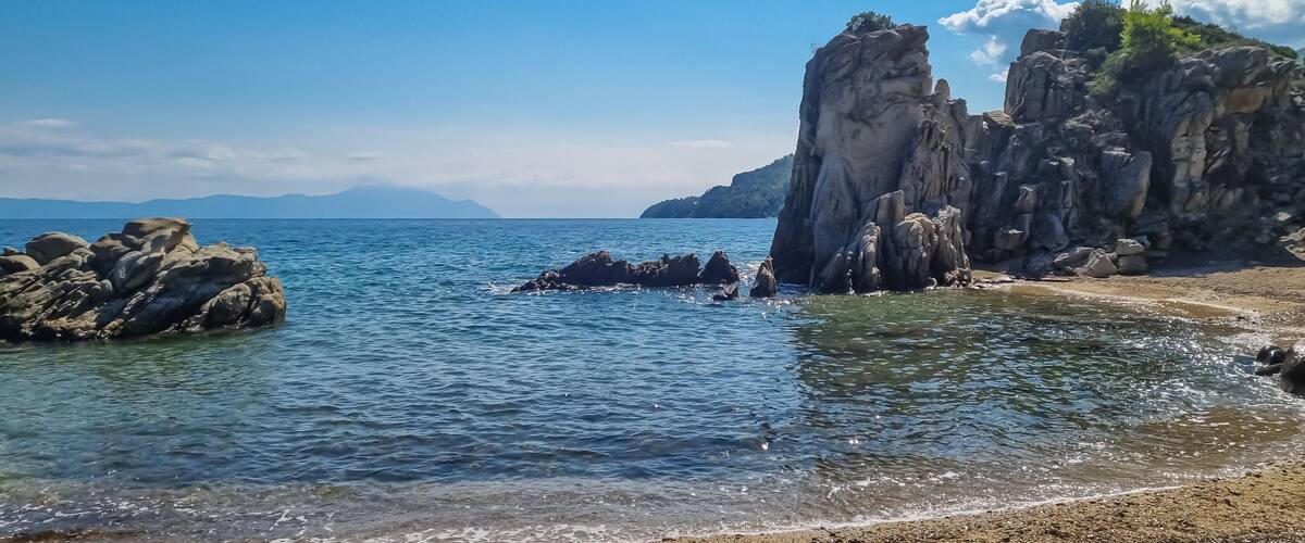 A coastal landscape with clear blue water, rocky formations, and distant mountains under a clear sky.