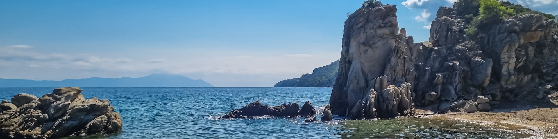 A coastal landscape with clear blue water, rocky formations, and distant mountains under a clear sky.
