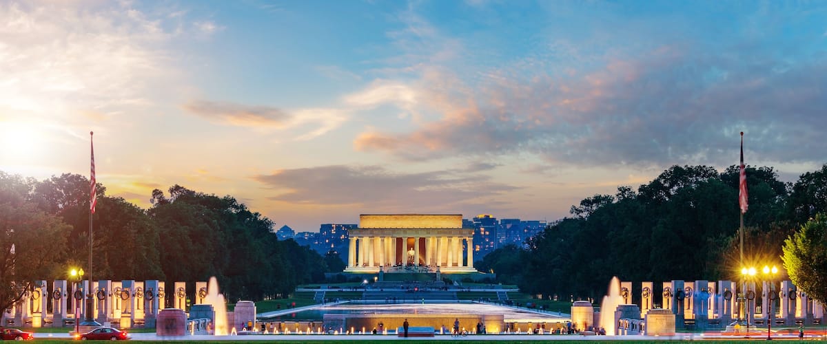 A large historical monument with a central statue, surrounded by a well-lit plaza and fountains, under a twilight sky.