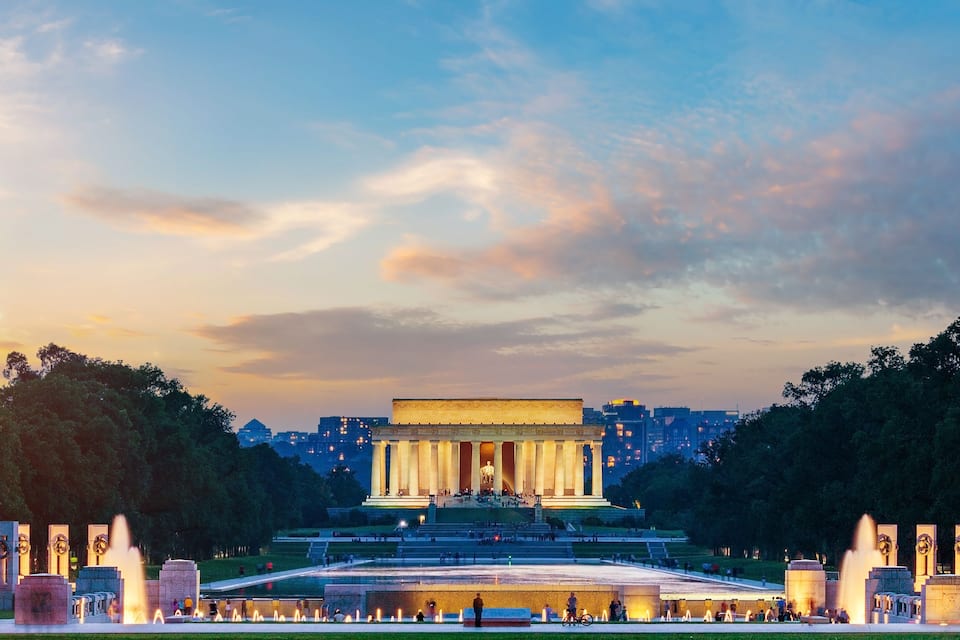 A large historical monument with a central statue, surrounded by a well-lit plaza and fountains, under a twilight sky.