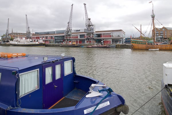 A harbor with a blue boat docked in the foreground, a large shipyard with cranes in the background, and a traditional wooden boat moored to the right.