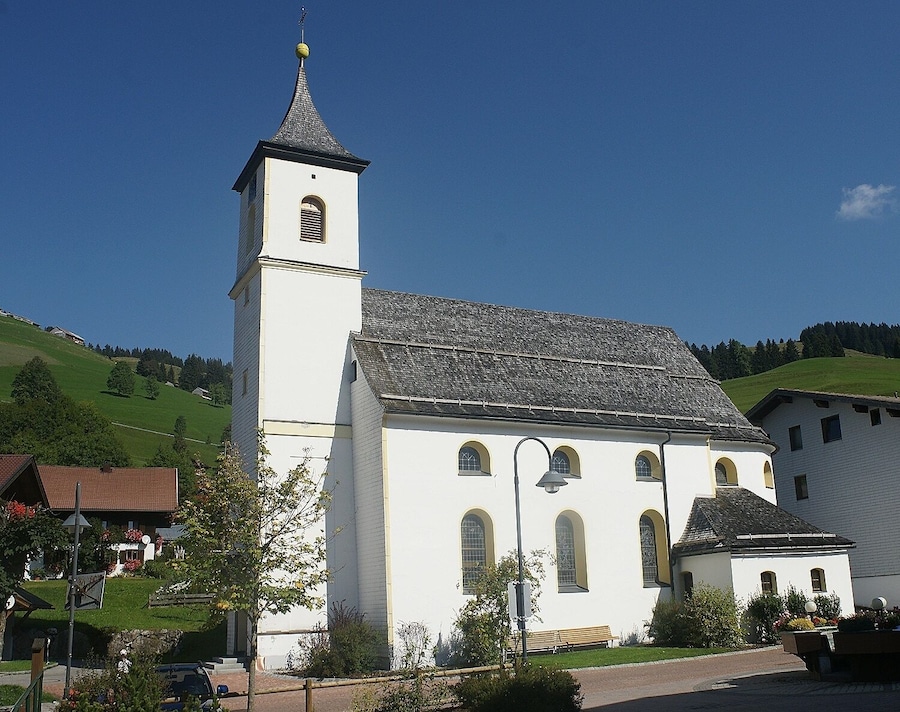 Eine Kirche mit hohem Glockenturm und steilem Dach, umgeben von Grünflächen und Häusern.
