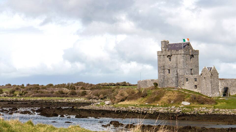 A historic stone castle with a tower, situated by a river with a grassy embankment and trees in the background.