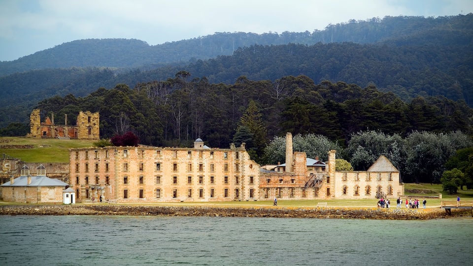 A historic stone building with a waterfront, surrounded by lush greenery and hills.