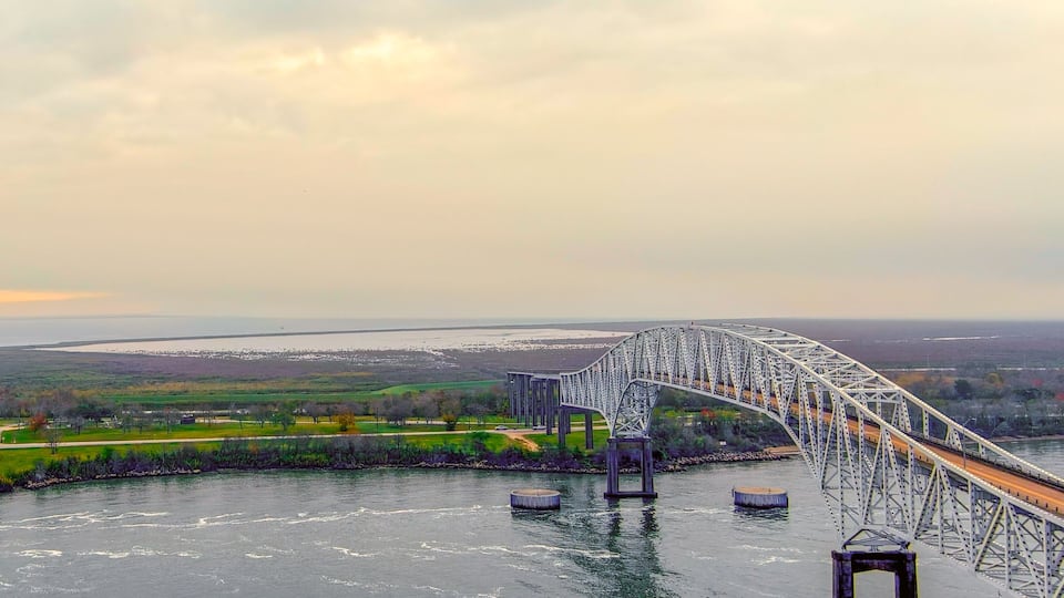 A large steel bridge spanning a river with a distant shoreline and fields.