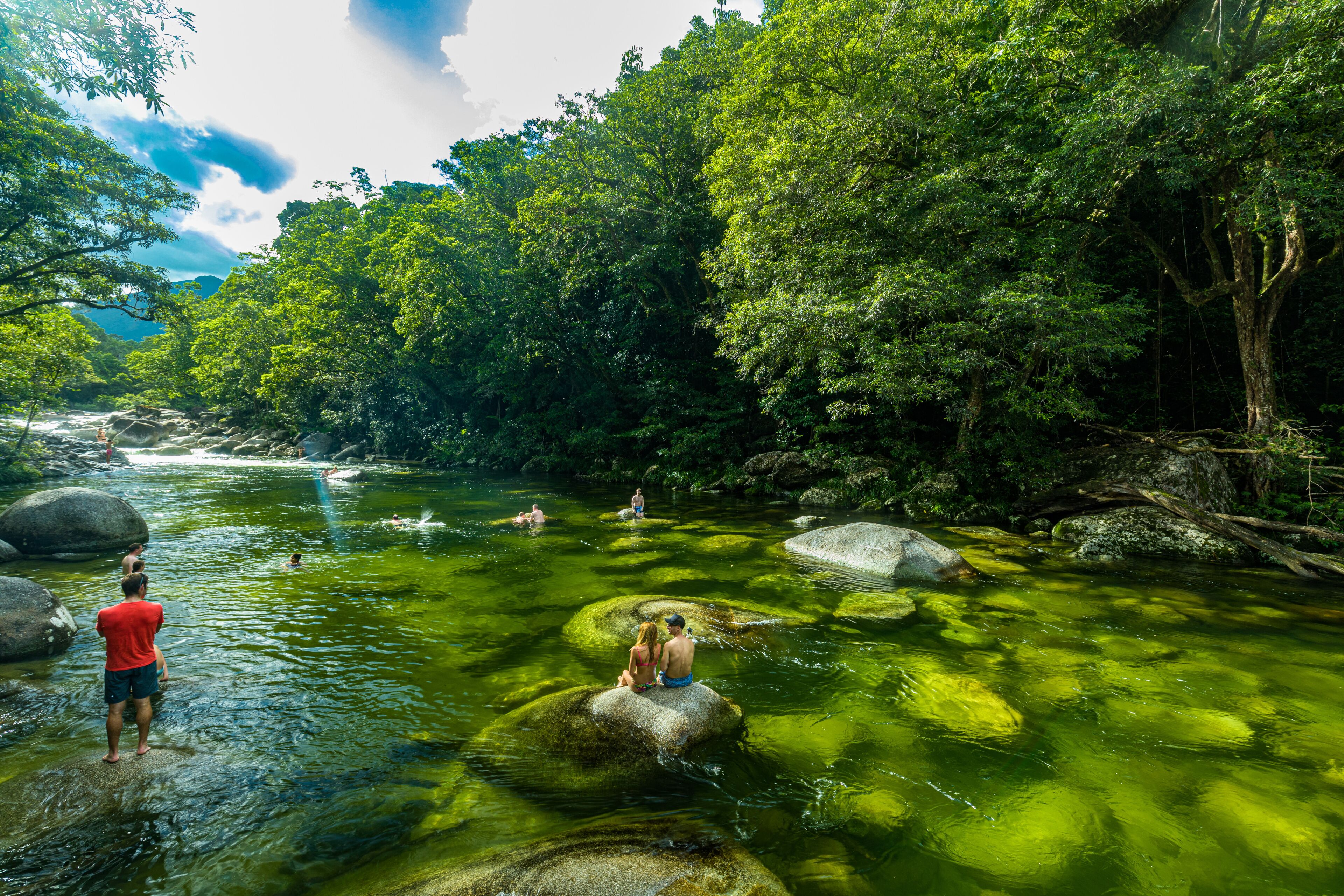 A river surrounded by dense green foliage, with people swimming and sitting on rocks.