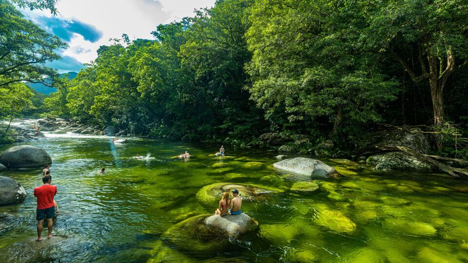 A river surrounded by dense green foliage, with people swimming and sitting on rocks.