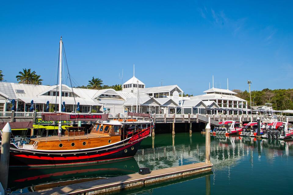 A marina with boats docked, a wooden boat with red and black stripes, and white buildings with a tropical backdrop.