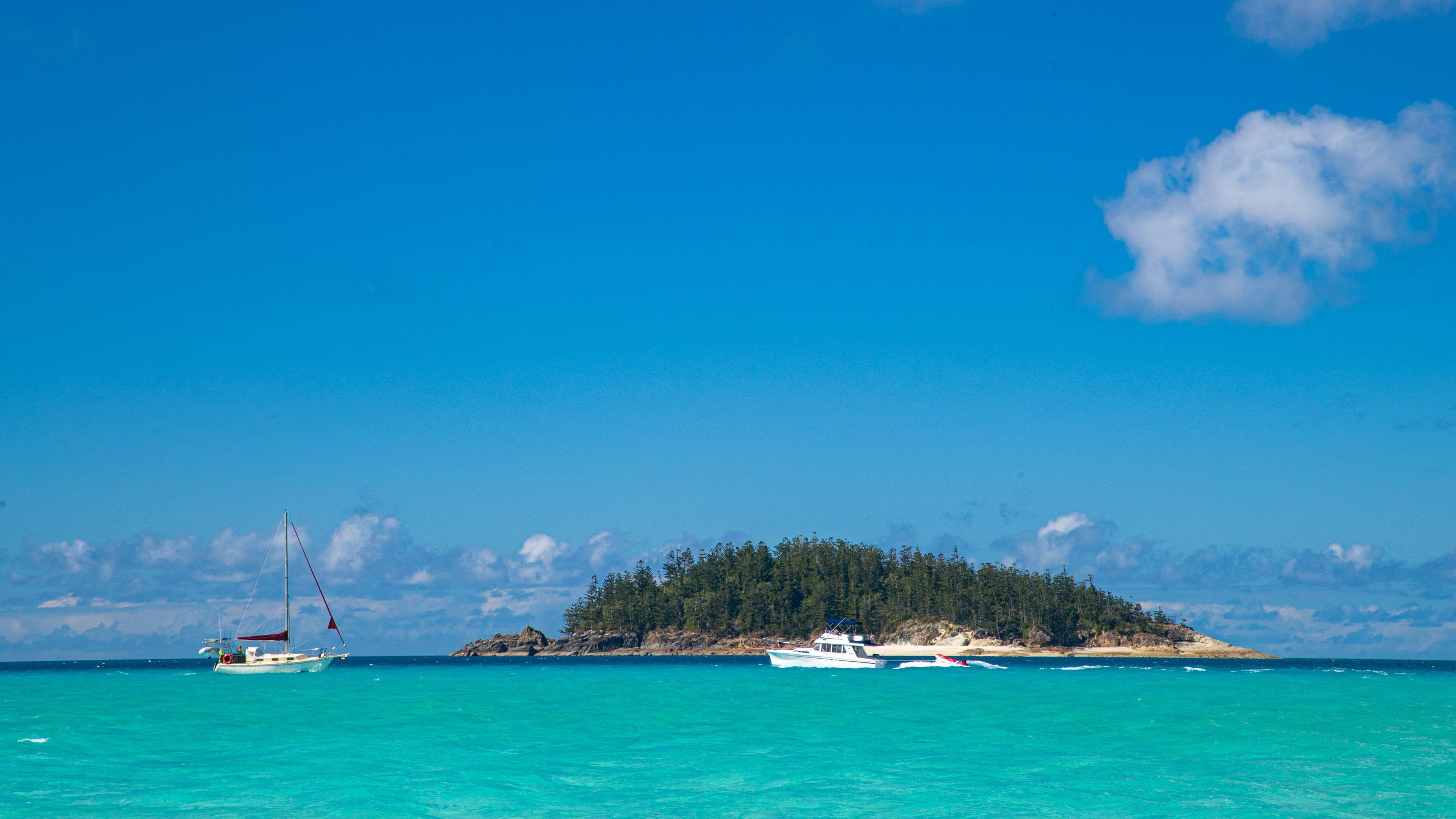 A turquoise sea with a sailboat and a motorboat near a small island with dense trees.