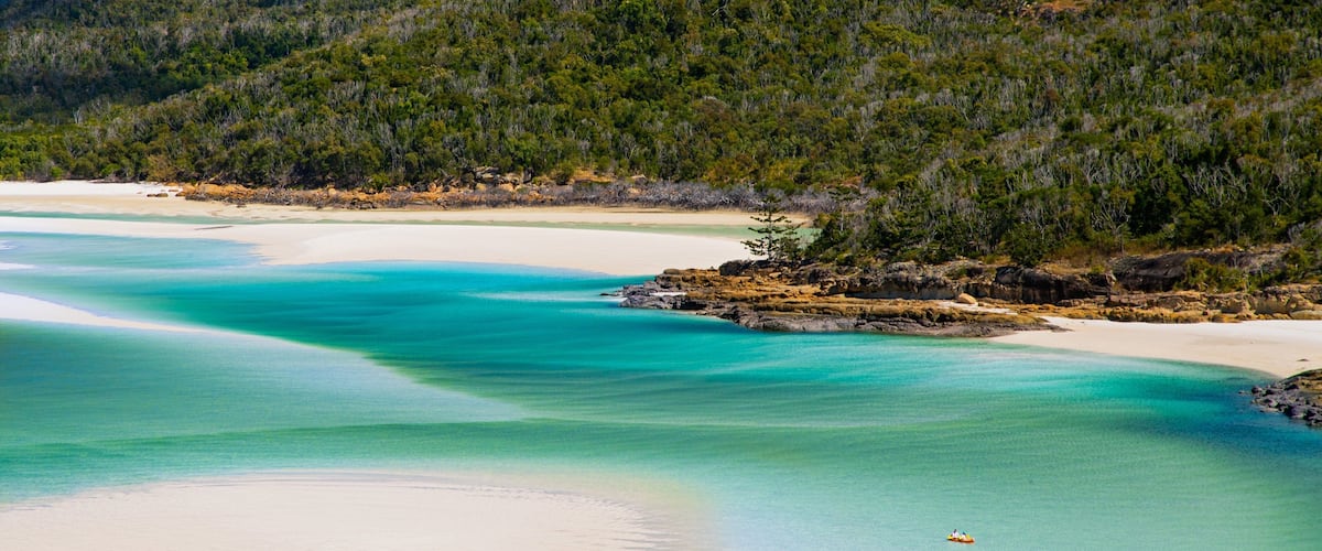 A coastal landscape with clear turquoise waters, white sandy beaches, and a forested hill in the background.