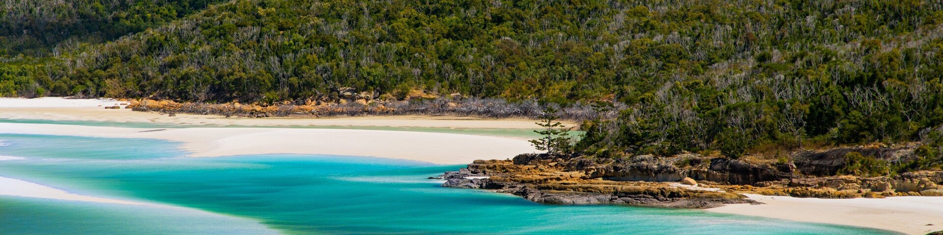 A coastal landscape with clear turquoise waters, white sandy beaches, and a forested hill in the background.