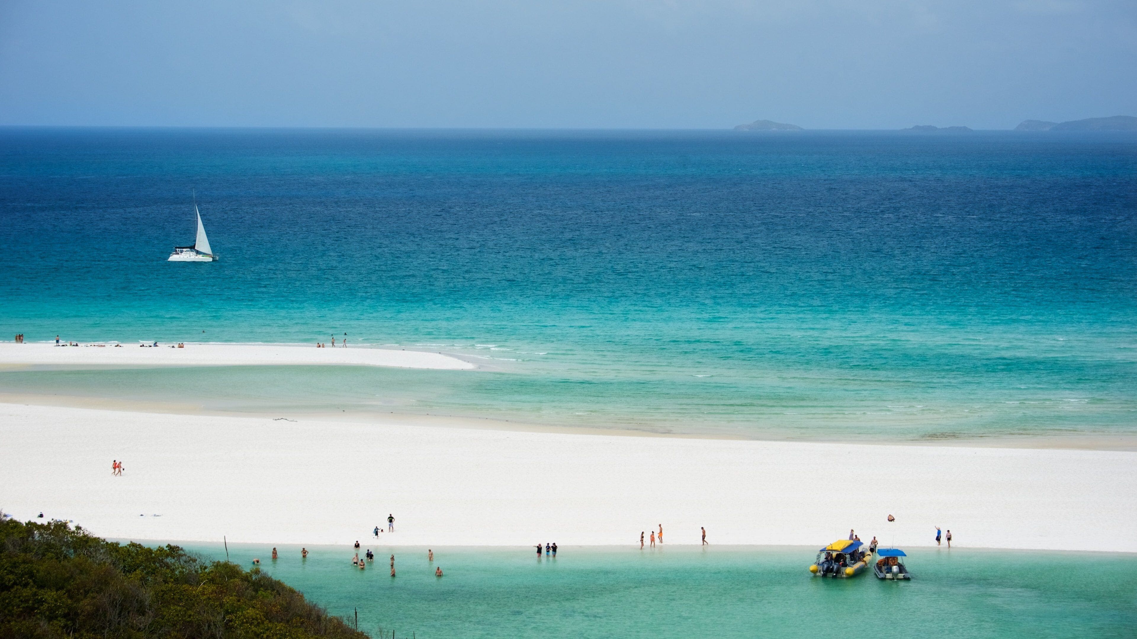 A beach with clear turquoise waters, a sailboat, and people swimming and sunbathing.