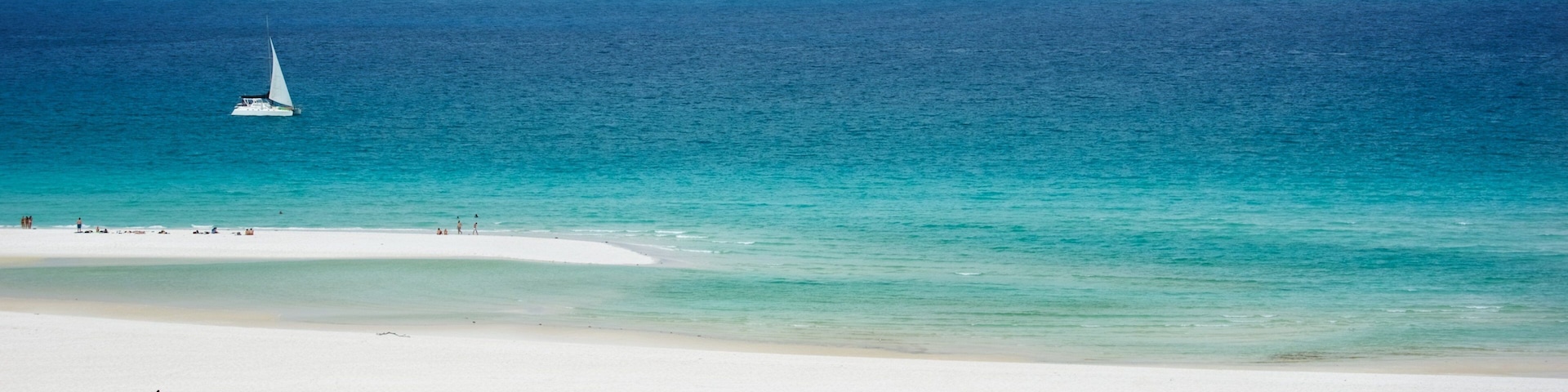 Uma praia com águas turquesa cristalinas, um veleiro e pessoas nadando e tomando sol.