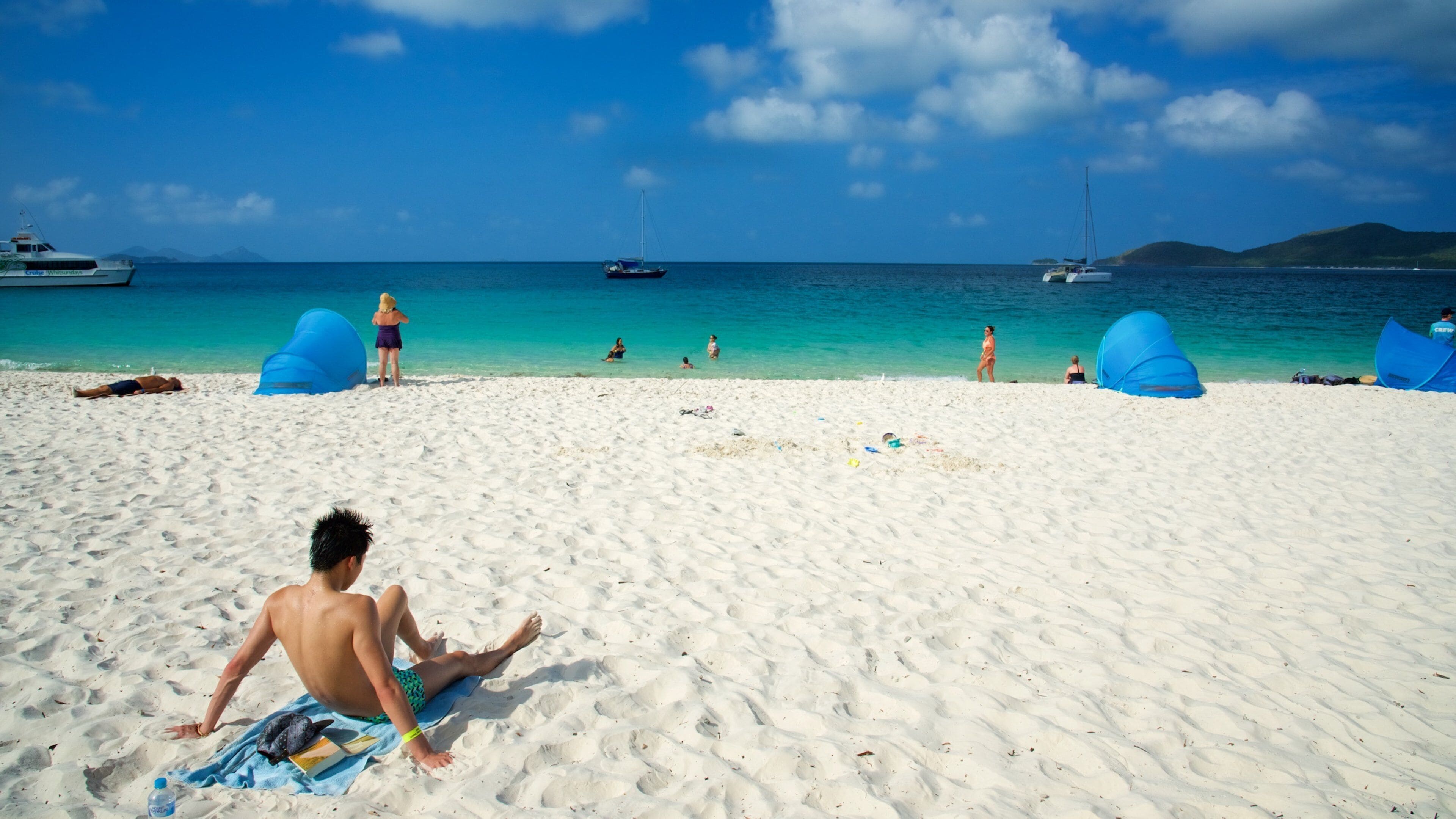 A beach with clear turquoise water, a man sitting on a towel, and boats in the distance.