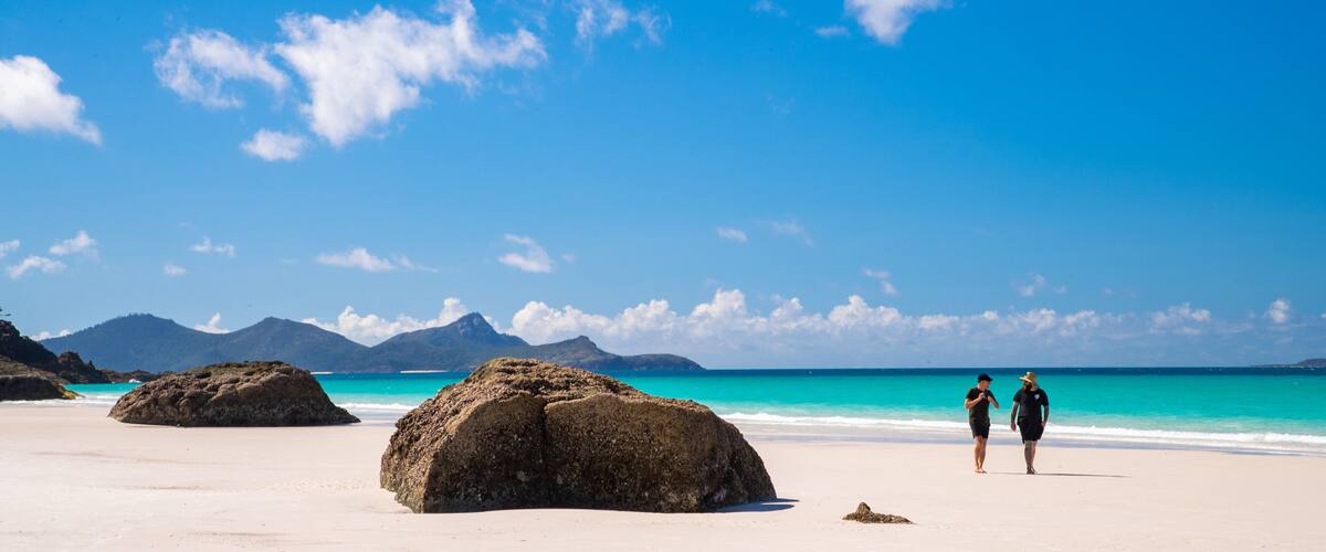 A beach with large rocks, clear turquoise water, and distant mountains under a blue sky.