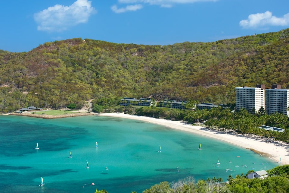 Uma área costeira com uma praia arenosa, águas turquesa cristalinas, veleiros e colinas verdes densas ao fundo.