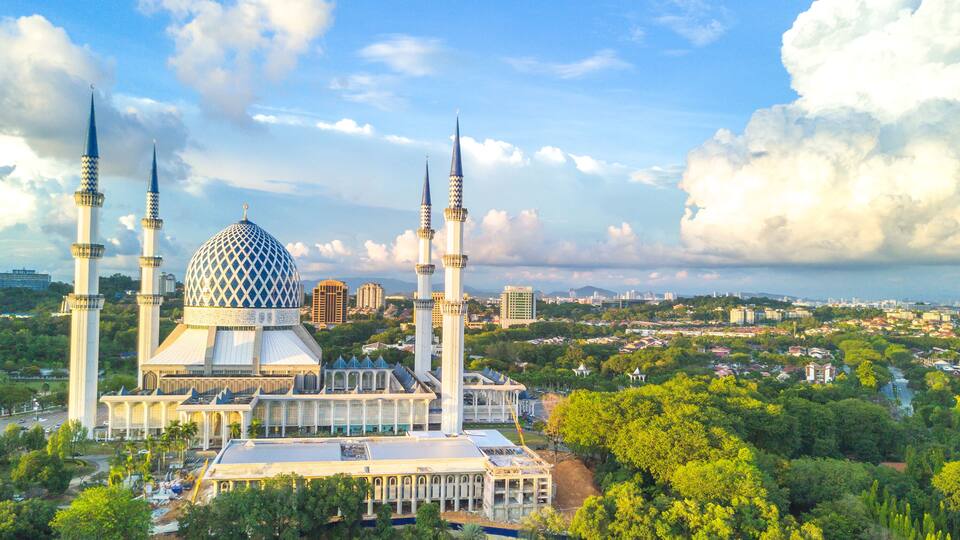A mosque with a large dome and four minarets, surrounded by greenery and a river.