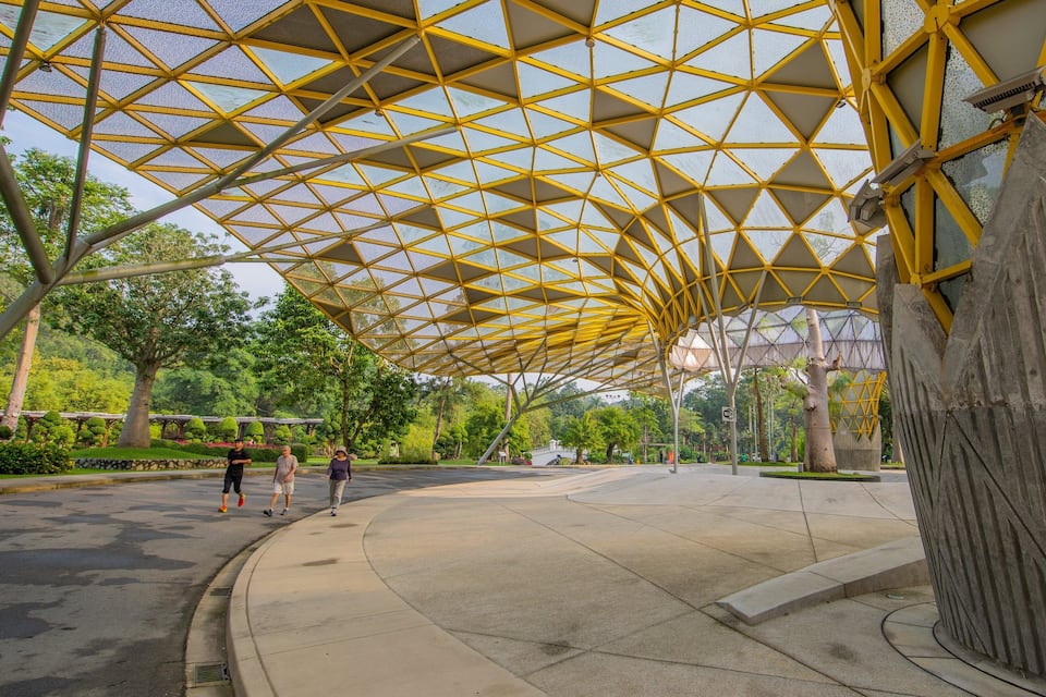 A covered walkway with a geometric yellow roof structure, surrounded by trees and a landscaped area.