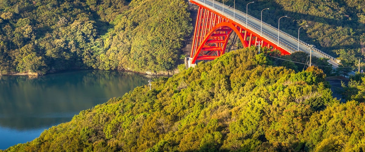 A red bridge spanning a river, surrounded by dense forest and hills.