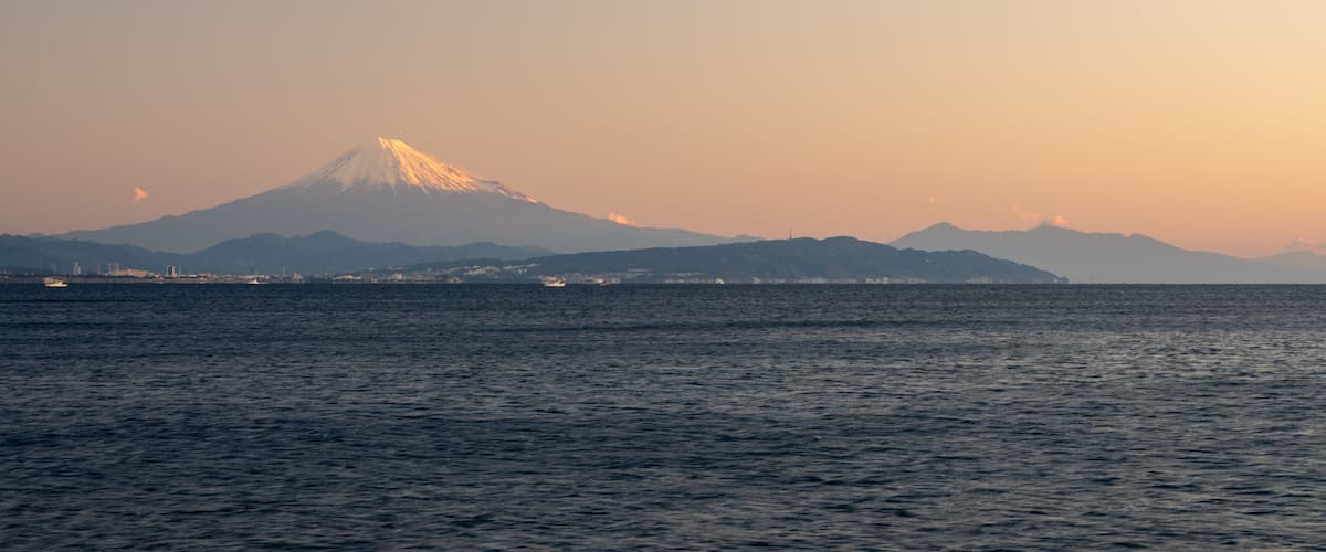 A mountain with a snow-capped peak, a body of water in the foreground, and a distant city skyline.
