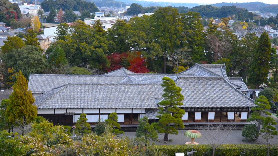 A traditional Japanese building with a tiled roof, surrounded by trees and a view of a town in the background.