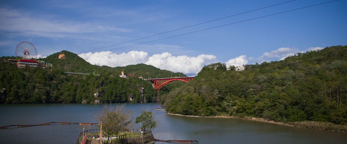 A red bridge over a river, surrounded by lush greenery and a Ferris wheel in the distance.