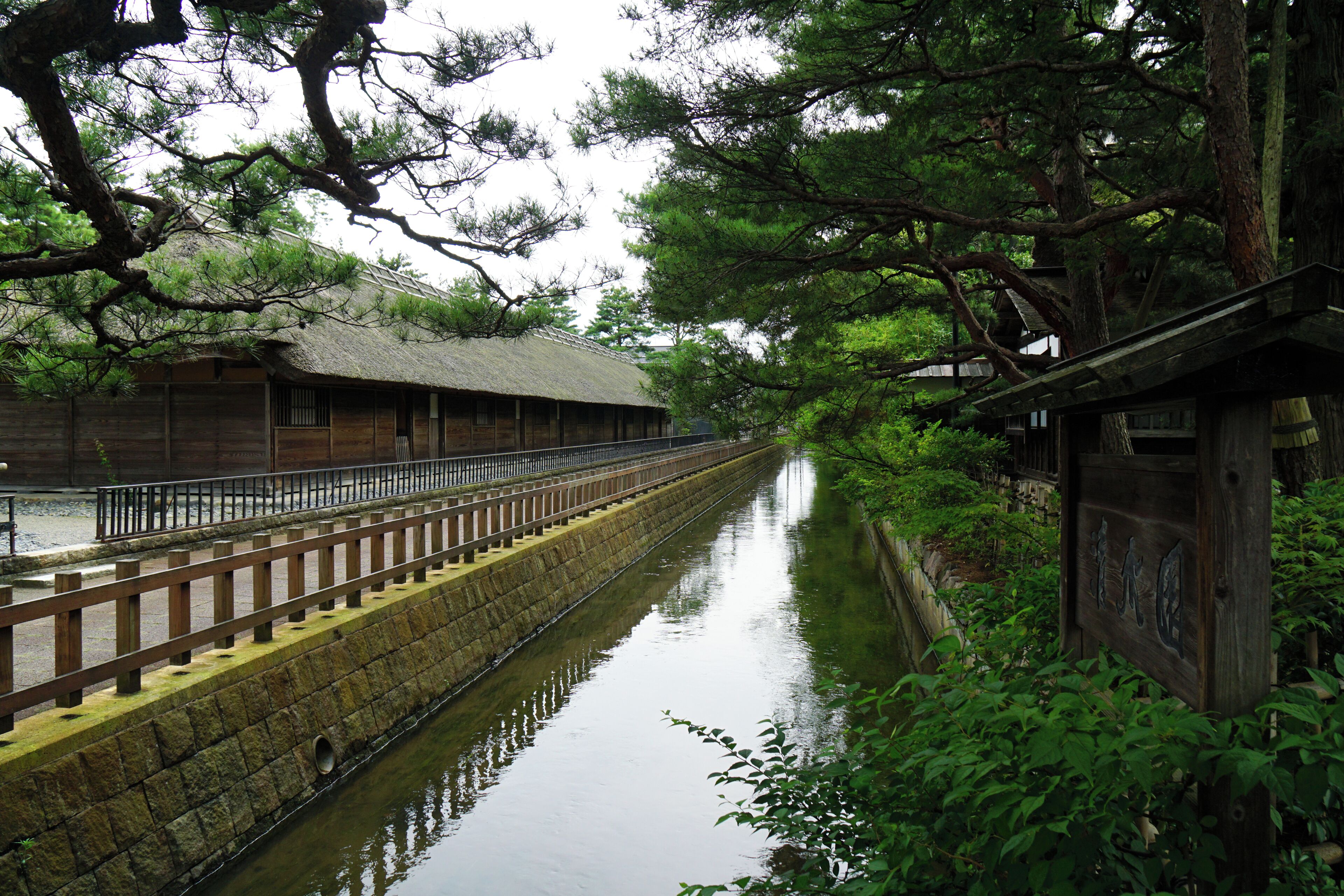 A traditional wooden building with a thatched roof, a stone wall, and a wooden bridge over a calm waterway.