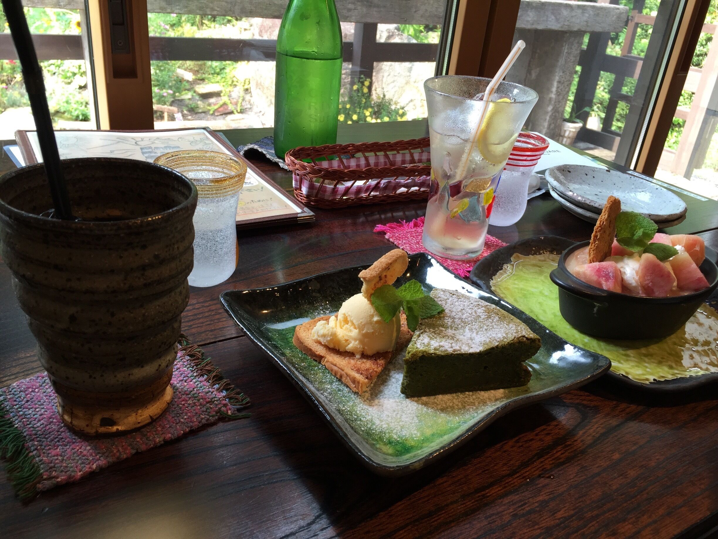 A table with a green bottle, a glass with a straw, a plate with ice cream and a slice of cake, and a bowl of fruit.
