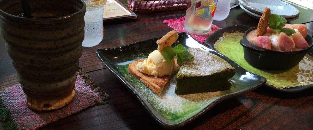 A table with a green bottle, a glass with a straw, a plate with ice cream and a slice of cake, and a bowl of fruit.