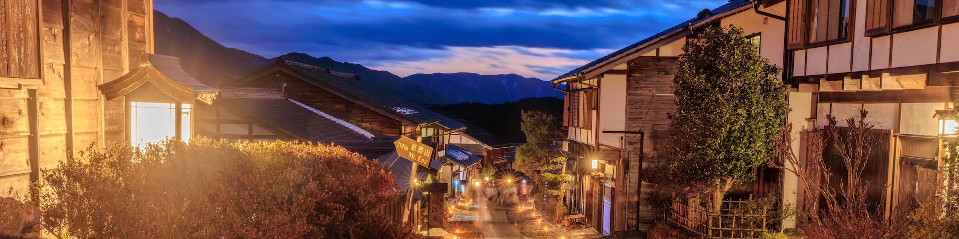 A stone pathway lined with traditional wooden buildings, illuminated by warm lights, leading towards a mountain range.