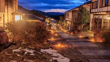 Uma trilha de pedra ladeada por edifícios de madeira tradicionais, iluminada por luzes quentes, que conduz a uma cordilheira.