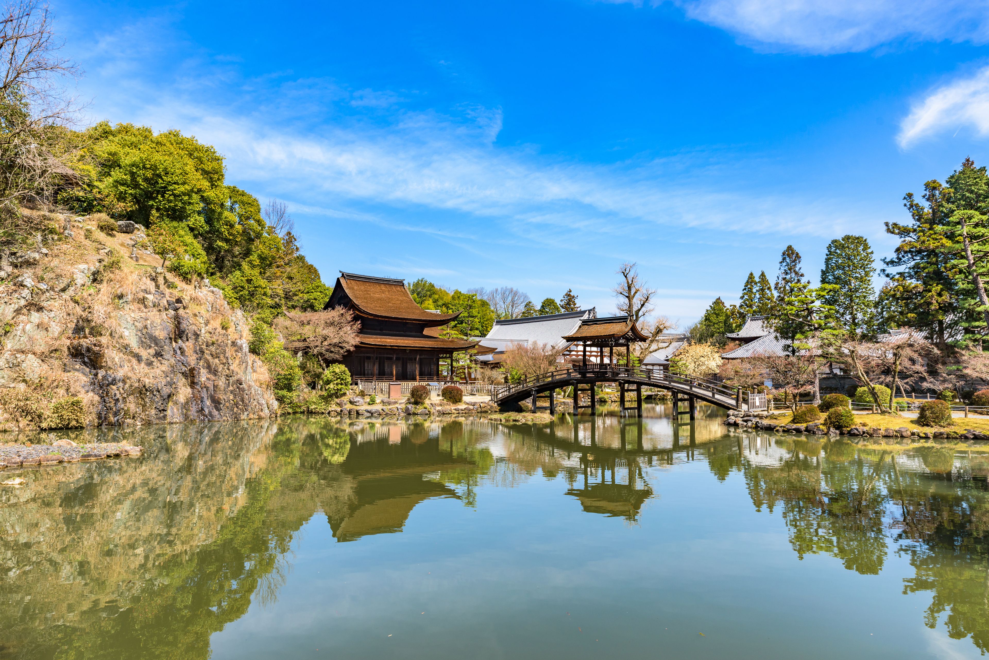 Un jardin japonais traditionnel avec un étang, un pont en bois et des bâtiments aux toits de chaume.