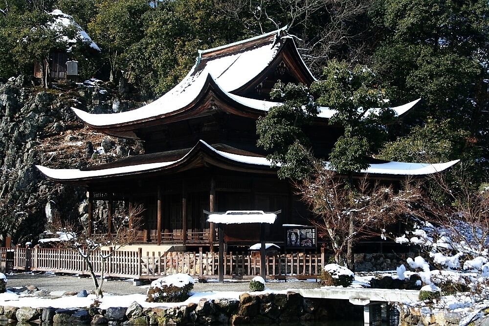 A traditional Japanese building with a snow-covered roof, surrounded by trees and rocks.