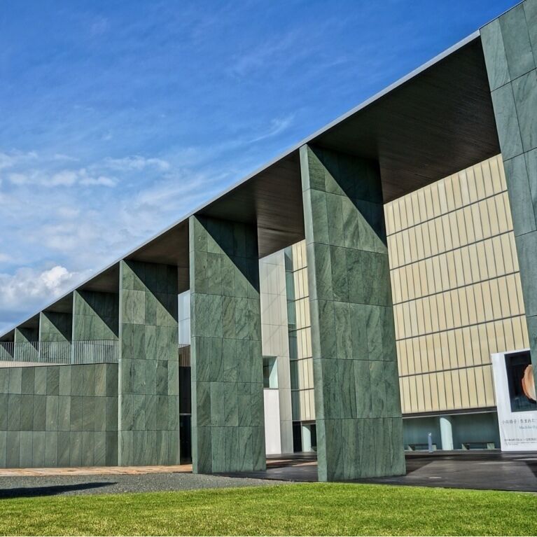 A modern building with green and beige panels, large columns, and a clear sky.