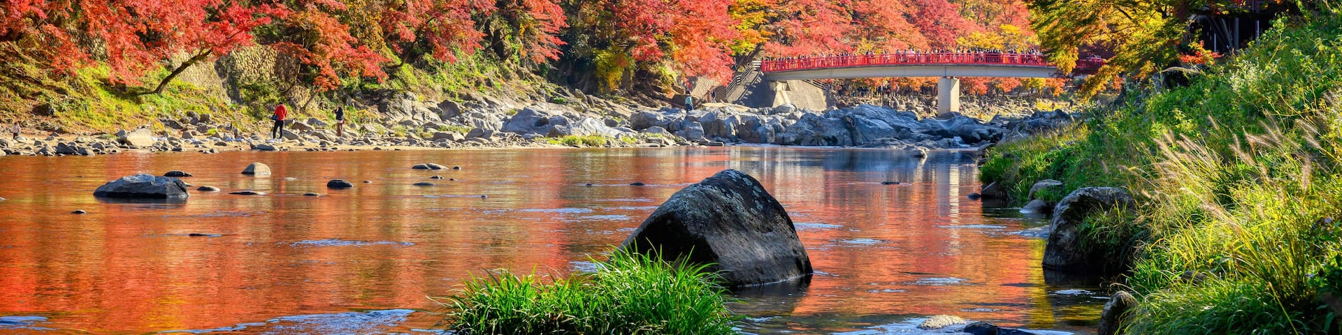 A river with autumn foliage, a bridge, and a mountainous backdrop.