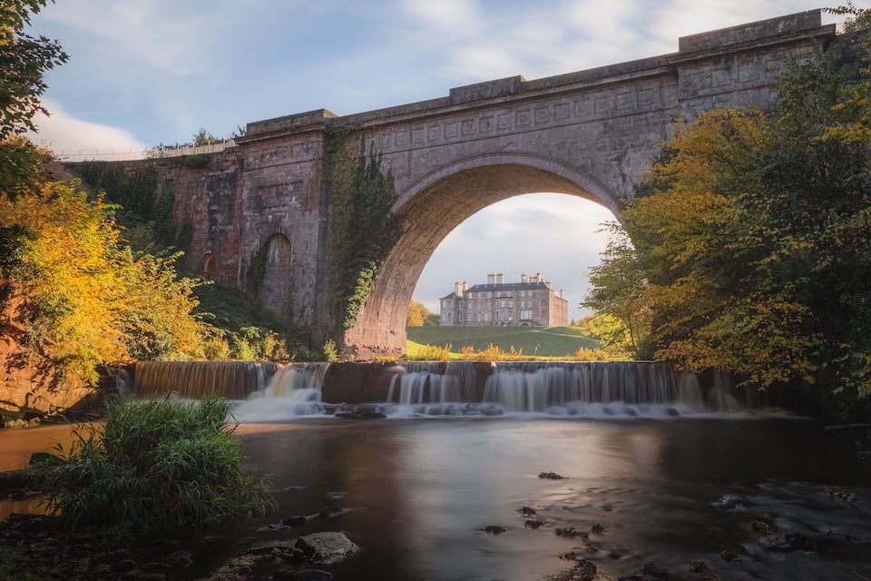 A stone bridge with an arch over a river, surrounded by trees and a castle in the background.