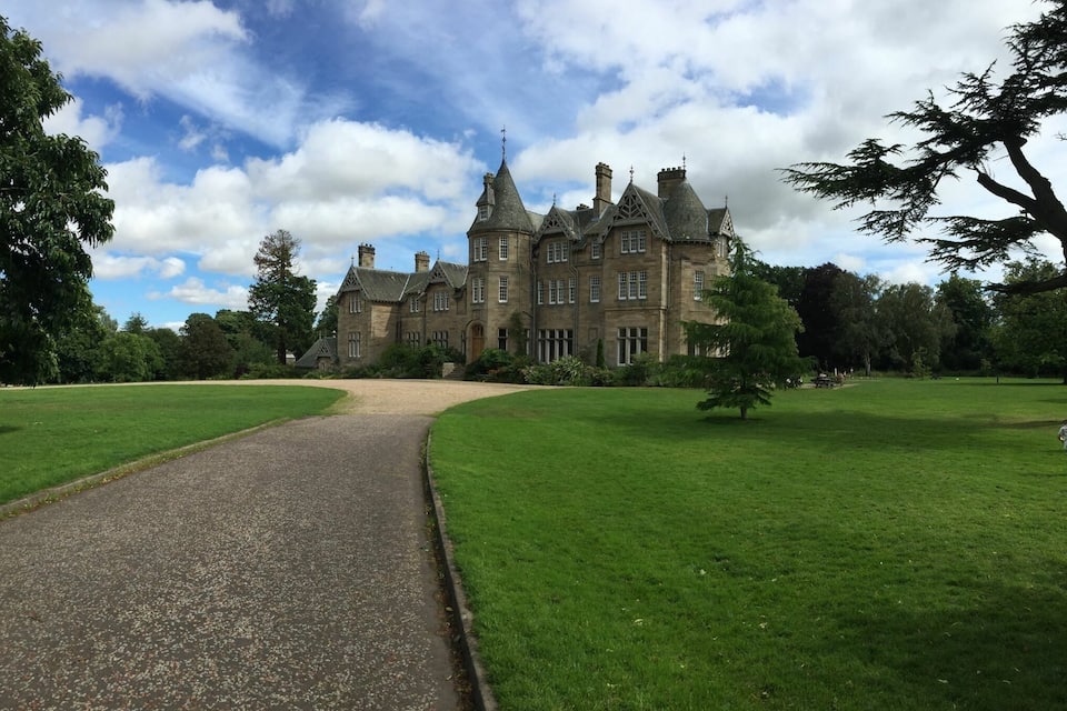 A large, historic building with a prominent tower and multiple chimneys, surrounded by lush green lawns and trees.